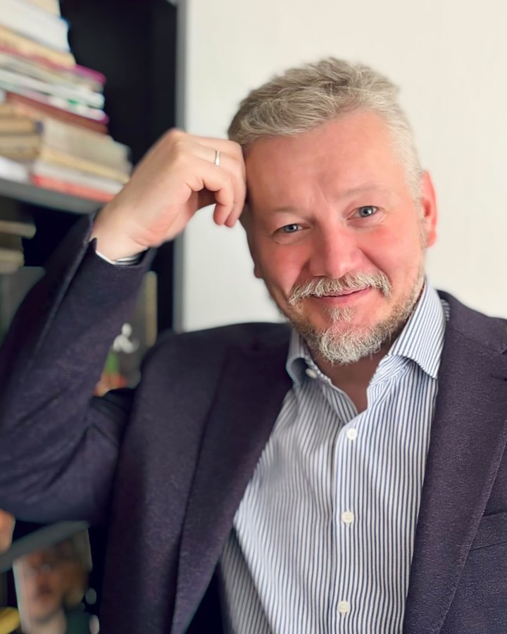 Maxim Romanov, seated in front of a bookshelf, in a dark jacket and striped shirt